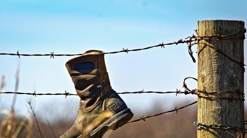 A rugged boot hanging on a rural barbed wire fence against a blue sky., tags: und - pexel